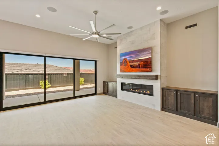 Spacious interior featuring a linear gas fireplace with a stacked stone surround, stained wood built-in cabinetry, wood-finish flooring, a contemporary multi-blade ceiling fan, and expansive sliding glass doors