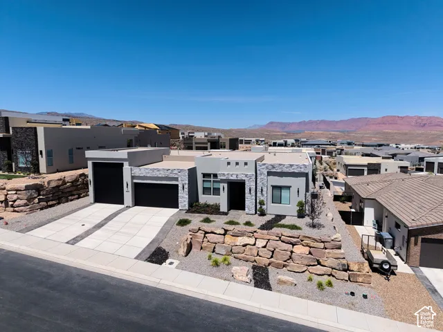 Contemporary stucco exterior with natural stone veneer accents, multi-car garage, concrete driveway, and tiered boulder retaining wall landscaping set against a mountainous desert backdrop