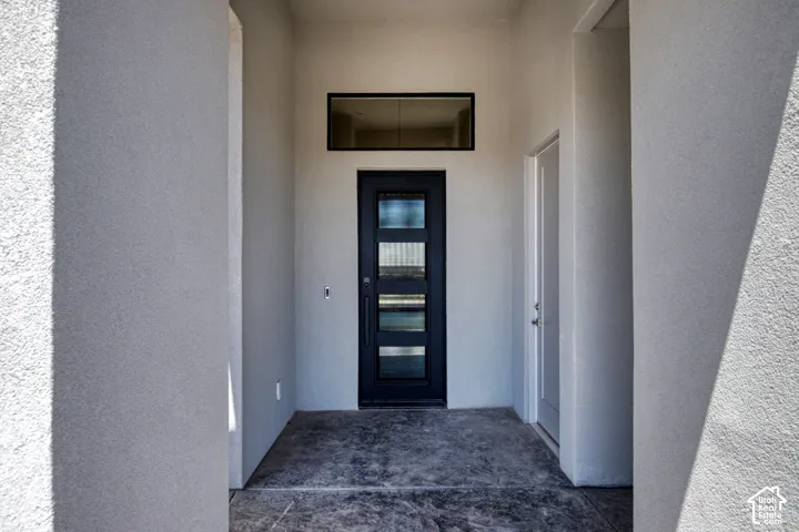 Covered entryway featuring smooth stucco walls, concrete flooring, a black front door with four glass panels, and a rectangular transom window