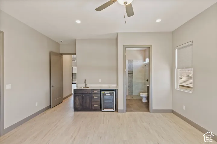Wet bar featuring dark-stained cabinetry, light-toned stone countertop, stainless steel sink, and built-in beverage refrigerator, complemented by light wood-finish flooring and a central ceiling fan