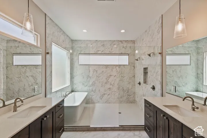 Dual vanities with stone countertops, freestanding soaking tub, walk-in shower with stone-patterned tile, glass shower enclosure, and pendant lighting