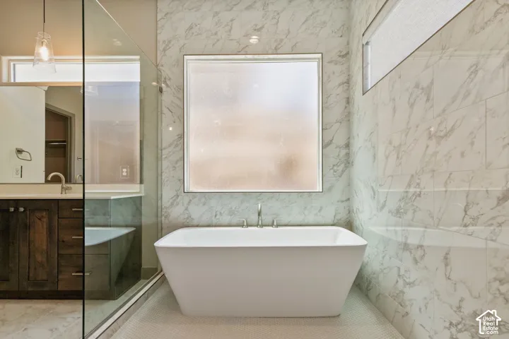 Freestanding soaking tub, floor-to-ceiling stone-finish tile surround, glass-enclosed shower, dark wood-finish cabinetry, and quartz countertop