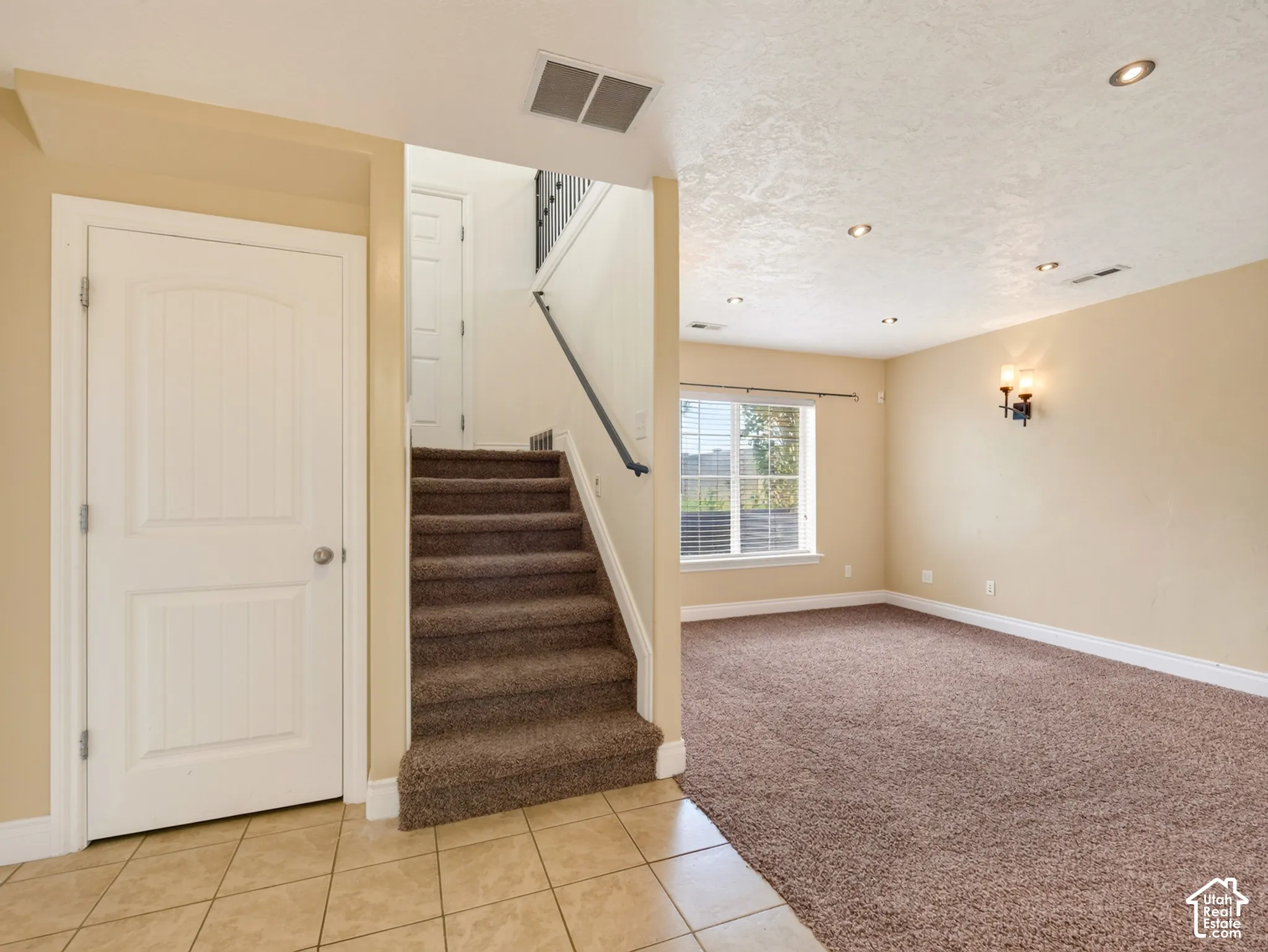 Interior space featuring tiled flooring, carpeted stairs with a dark metal handrail, recessed ceiling lighting, wall-mounted sconce lighting, and a window with a grid pattern