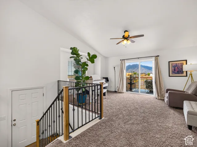 Vaulted ceilings, carpeted flooring, wrought iron stair railing with wood newel posts, ceiling fan with light fixture, and sliding glass door with mountain scenery
