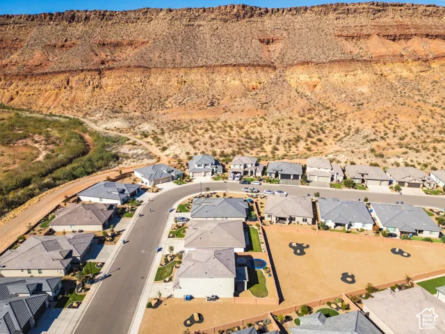 Aerial perspective of a residential neighborhood featuring paved streets, single-story homes with pitched roofs, manicured lawn segments, and a prominent sandstone cliff backdrop