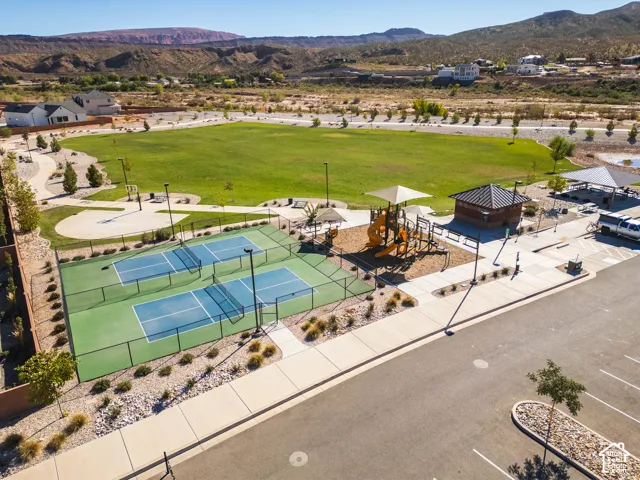 Community common area featuring two fenced pickleball courts, a playground with shade structures, a covered pavilion, and expansive open grass space set against a mountain backdrop