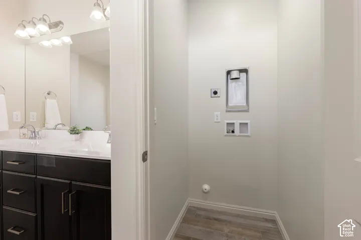 Dark-toned cabinetry, light-toned countertop with dual integrated sinks, wide wall-mounted mirror, polished chrome fixtures, and dedicated laundry hookups with wood-finish flooring