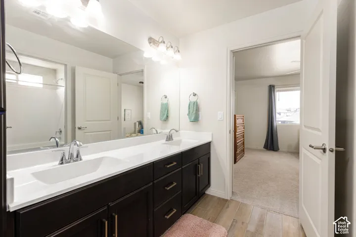 Bathroom featuring a dual-sink vanity with white countertops, dark cabinetry, wood-finish flooring, and a full-width mirror