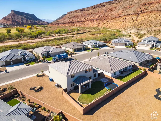 Aerial perspective of a multi-story residence featuring a covered patio, a fenced backyard with landscaped turf, a two-car garage, and desert topography with mountain backdrops