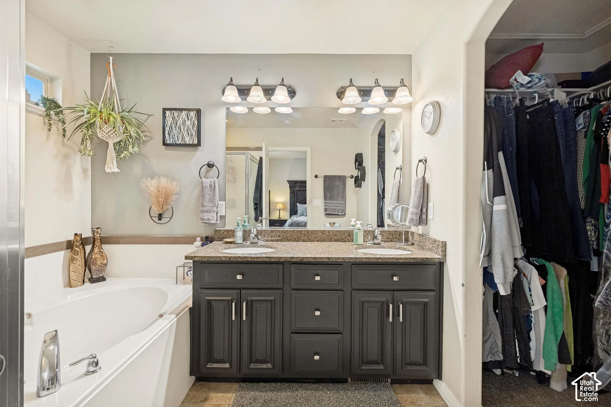 Dual-sink vanity with dark cabinetry, stone-finish countertop, full-width mirror, and deep soaking tub with chrome fixtures