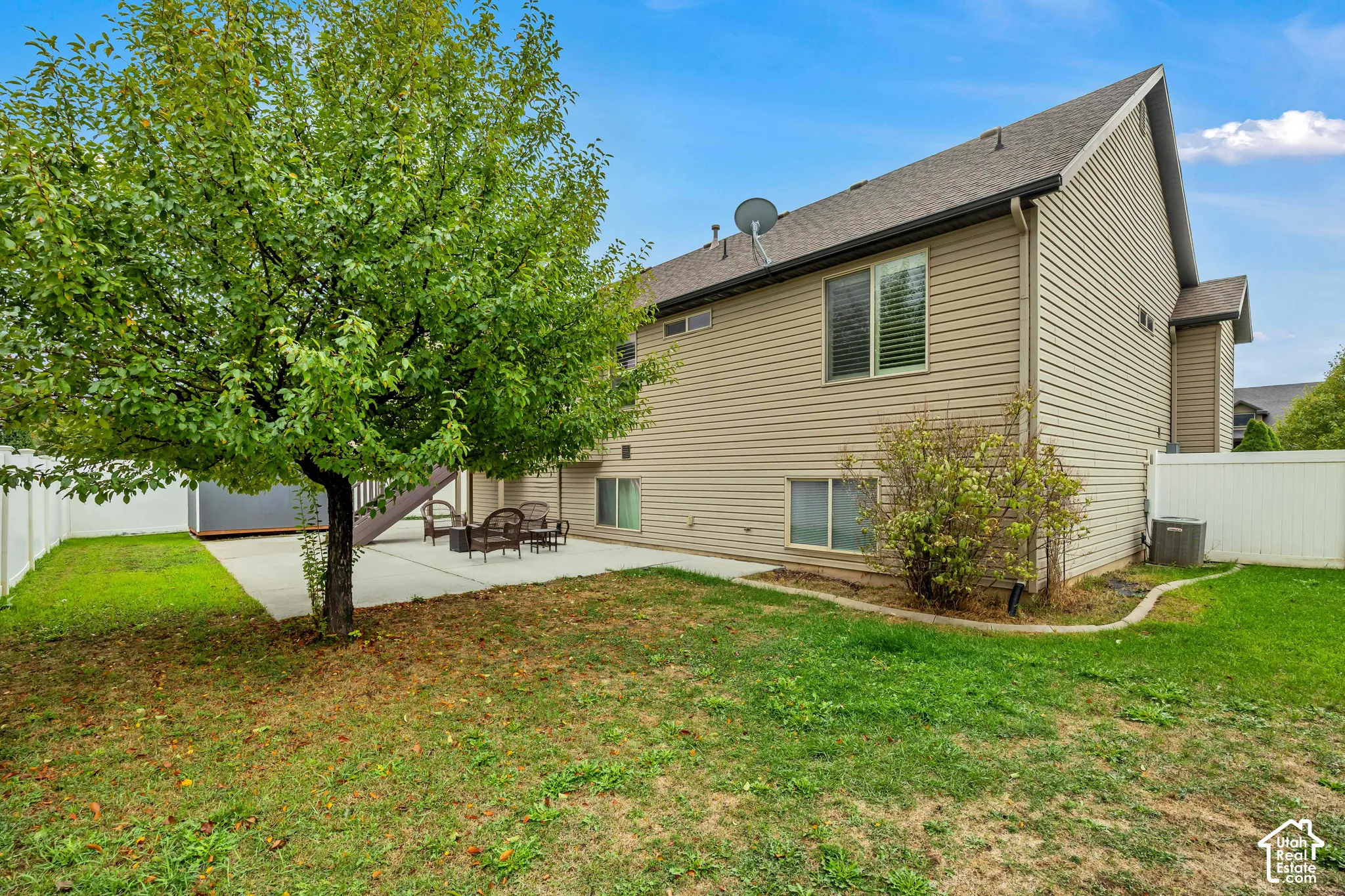 Rear exterior featuring tan vinyl siding, a concrete patio, white vinyl privacy fencing, and a mature tree