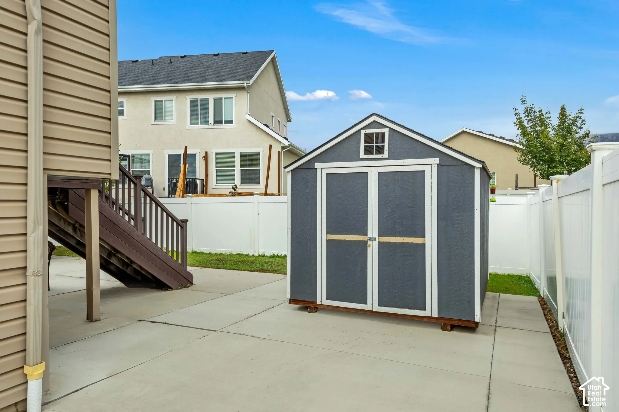 Exterior storage shed featuring a gabled roof and double-door entry, situated on a concrete patio enclosed by white vinyl fencing