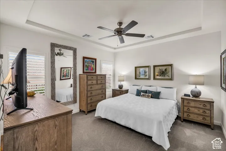 Bedroom featuring a tray ceiling, ceiling fan with light fixture, neutral wall-to-wall carpeting, and plantation shutters