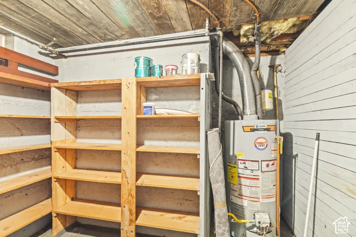 Utility area featuring built-in wooden shelving, painted masonry walls, exposed wood ceiling joists, and a direct-vent water heater