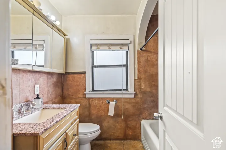 Bathroom featuring natural stone-finish wall tile, wooden vanity with a stone-finish countertop, undermount sink, tri-fold medicine cabinet, and an arched shower alcove