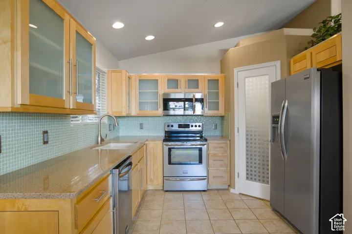 Kitchen featuring natural wood cabinetry with frosted glass inserts, stone countertops, light-toned tiled backsplash, stainless steel appliances, and recessed lighting
