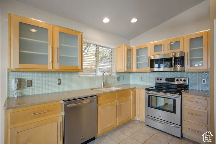 Kitchen featuring light-tone wood cabinetry with frosted glass inserts, stainless steel appliances, a tile backsplash, stone-finish countertops, and recessed lighting