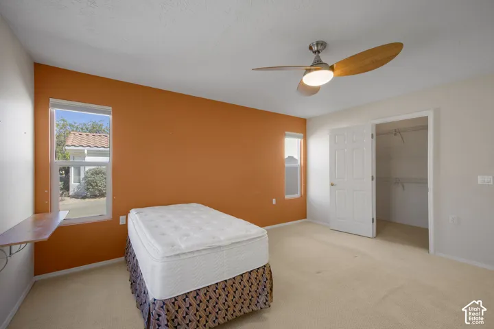 Room featuring neutral wall-to-wall carpeting, dual-pane windows, a wall-mounted floating shelf, a ceiling fan with integrated lighting, and a walk-in closet with double-door entry