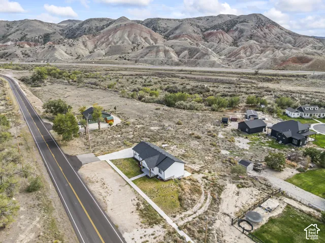 Aerial view of property featuring a white exterior residence, dark-toned roof, concrete driveway, and expansive natural landscape backdropped by mountain terrain