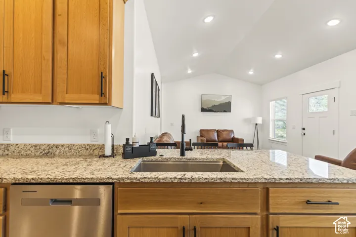Kitchen featuring natural-finish wood cabinetry, stone countertops, stainless steel undermount sink with matte black faucet, integrated stainless steel dishwasher, and vaulted ceilings