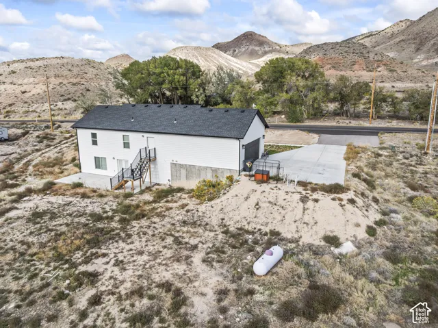 Two-story residential property featuring a white exterior, dark shingle roof, concrete driveway, and metal exterior staircase, set against a backdrop of mountainous terrain