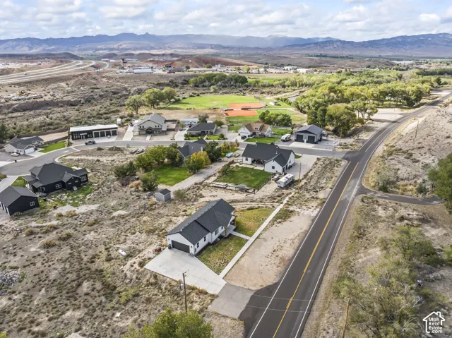 Aerial perspective of a single-story residence featuring a dark shingle roof, white siding, attached garage, concrete driveway, and prominent mountain horizon