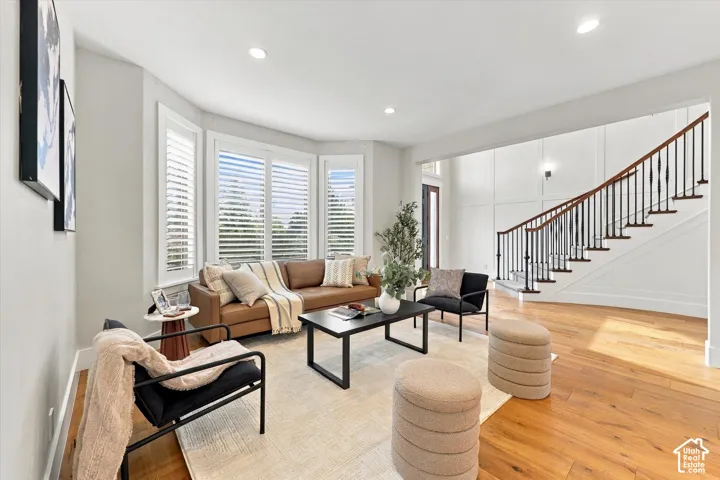 Living space featuring wood-finish flooring, a three-panel bay window with shutter-style blinds, recessed lighting, and a staircase with a wood handrail and iron balusters