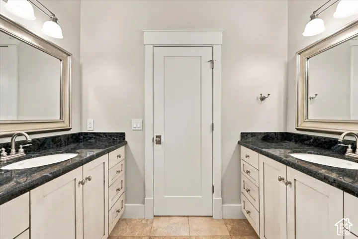 Dual vanity featuring stone countertops, undermount sinks, custom cabinetry, framed mirrors with integrated lighting, and tile flooring