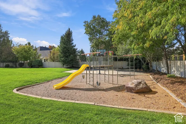 Expansive backyard featuring a large grass lawn, perimeter vinyl fencing, mature trees, and a dedicated play area with a playground structure and basketball hoop