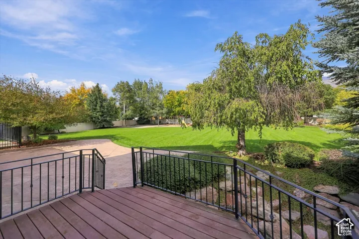 Expansive backyard featuring a wood-finish deck, wrought-iron railing, stamped concrete patio, manicured lawn, and mature trees