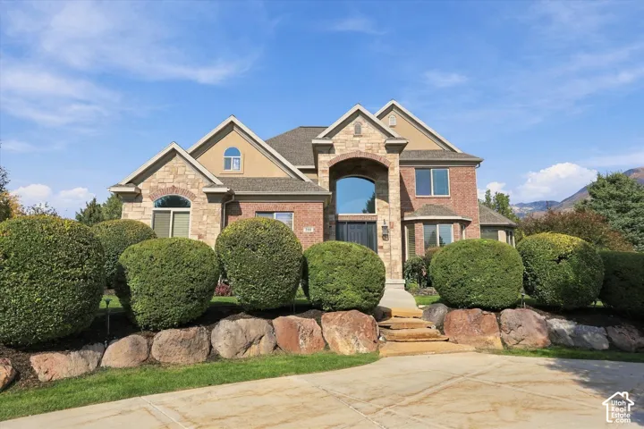 Two-story exterior featuring a brick and stone facade, arched entryway, multi-gabled roofline, stacked-stone retaining wall, and mountain backdrop