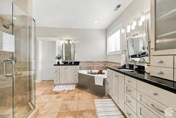 Bathroom featuring a glass-enclosed shower, corner soaking tub with metallic tile surround, dual separate vanities with dark countertops, and tile flooring