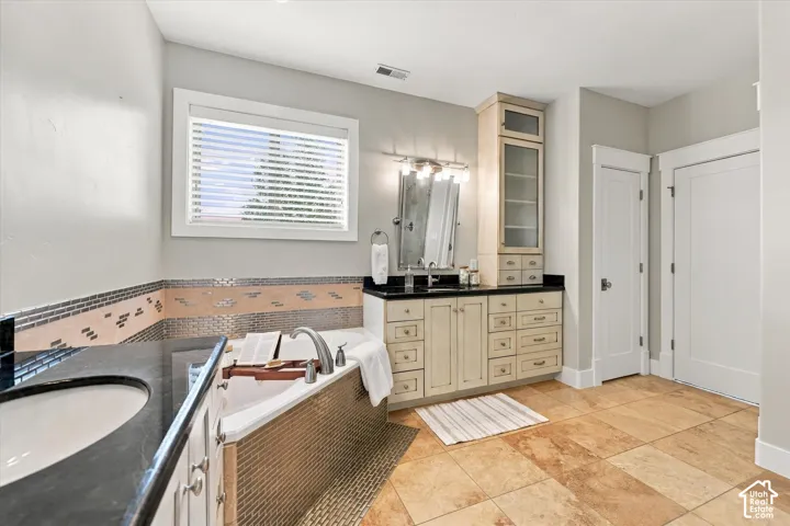 Bathroom featuring a custom built-in linen tower, vanity with dark stone countertop, corner jetted tub with metallic tile surround, and stone-finish floor tile