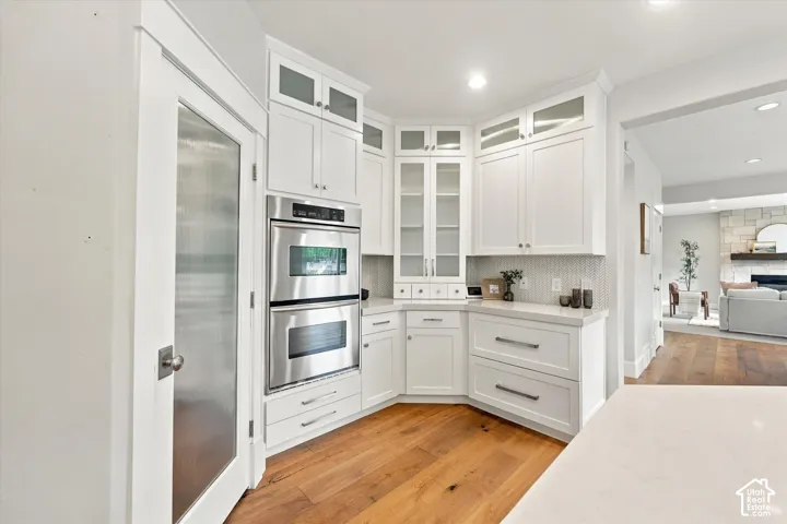 Kitchen featuring white shaker-style cabinetry with glass-front uppers, stainless steel double wall ovens, stone-composite countertops, and wood-finish flooring
