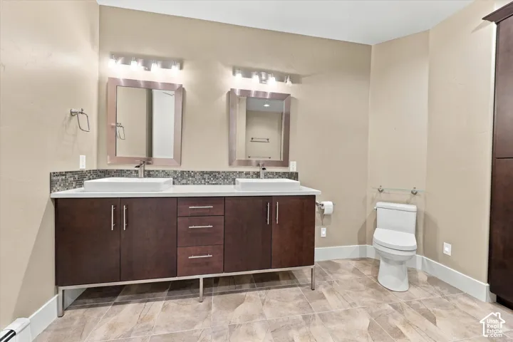 Dual-sink vanity with dark wood-finish cabinetry, rectangular vessel sinks, light-toned countertops, mosaic tile backsplash, and two wall-mounted metallic light fixtures