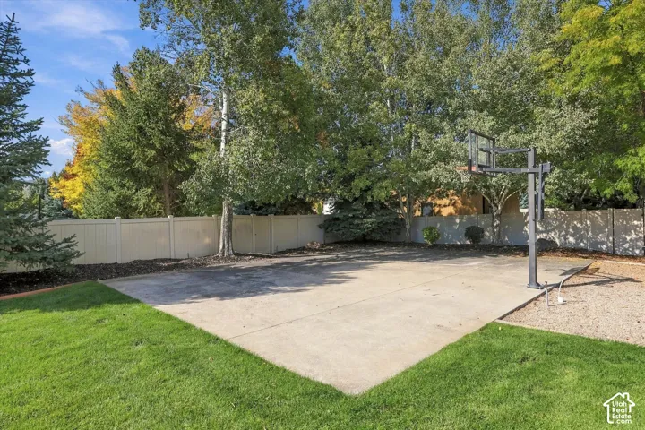 Concrete sport court featuring a built-in basketball hoop, enclosed by solid vinyl fencing and surrounded by a manicured lawn and mature landscaping
