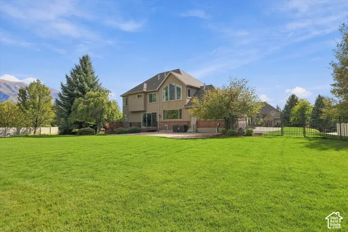 Expansive backyard featuring a manicured lawn, paved patio, mature trees, and perimeter metal fencing