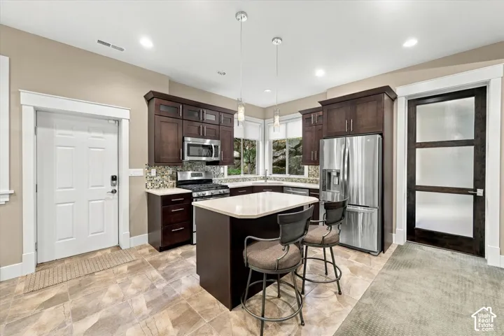 Kitchen featuring dark wood cabinetry, a central island with a solid surface countertop, stainless steel appliances, mosaic tile backsplash, and a three-panel frosted glass door