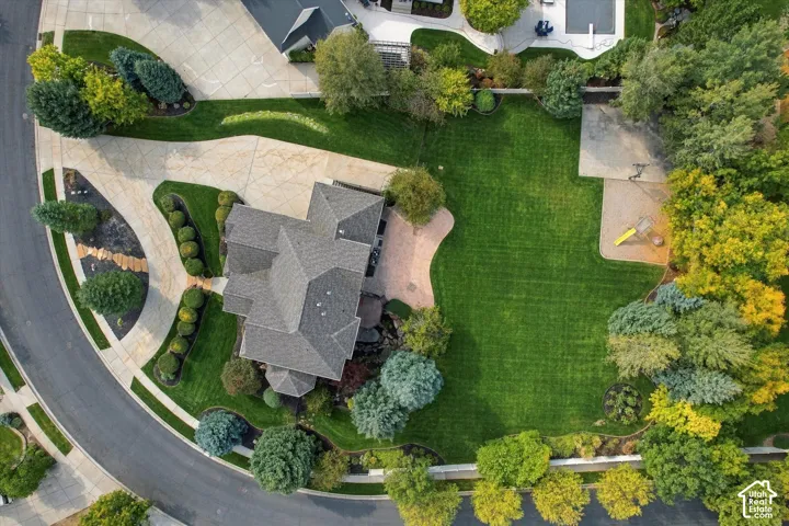 Aerial view of property featuring a multi-pitched shingle roof, expansive manicured lawn, stone-paved patio, extensive mature landscaping, and curved concrete driveway