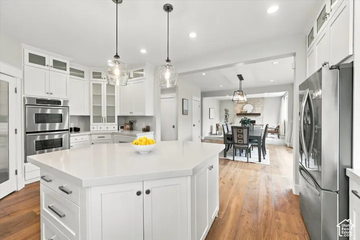 Kitchen featuring white cabinetry with glass-front uppers, a central island with a quartz countertop, wood-finish flooring, built-in stainless steel double ovens, and pendant lighting