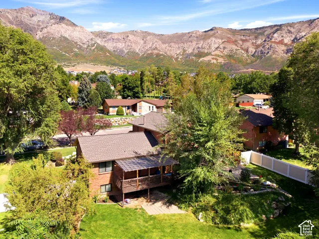 Brick exterior, elevated wooden deck with overhead pergola, expansive lawn, stone-accented tiered landscaping, and prominent mountain backdrop