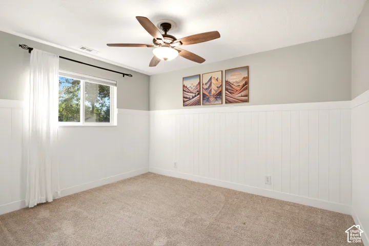 Neutral-toned interior featuring white vertical wall paneling, carpeting, a ceiling fan with integrated lighting, and a single sliding window