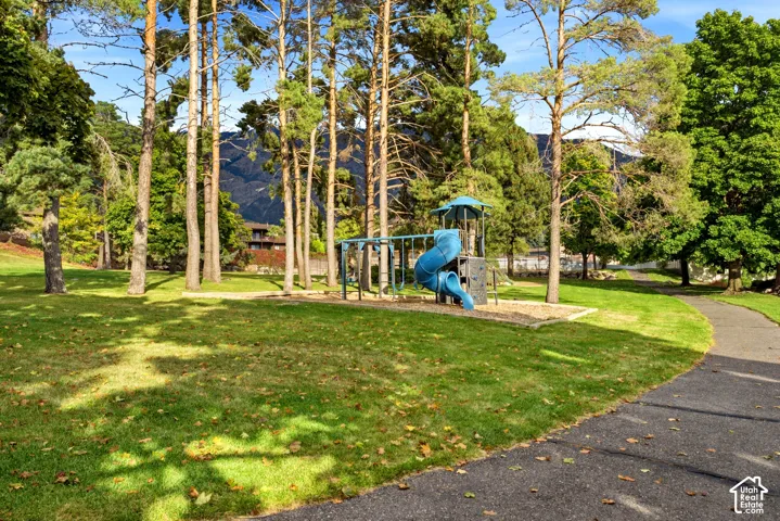 Expansive grounds featuring a playground structure, mature coniferous trees, a manicured lawn, and an asphalt walking path set against a mountain backdrop
