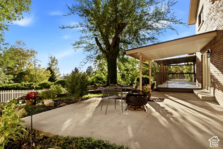 Concrete patio featuring a covered deck, brick exterior siding, mature trees, and a picket fence