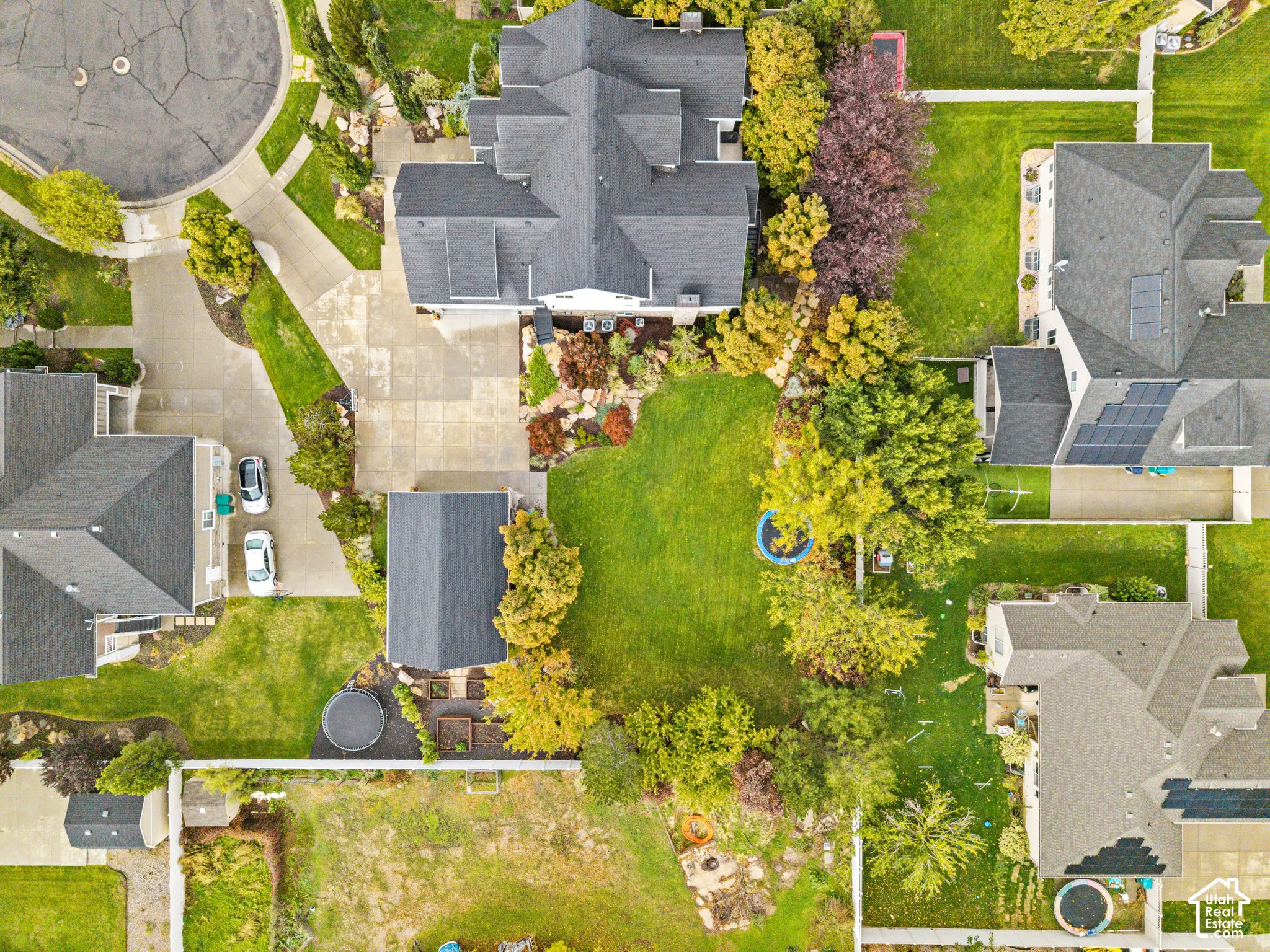 Aerial view of a residential property featuring a large backyard, concrete driveway, multi-gabled roofline, established trees, and perimeter fencing