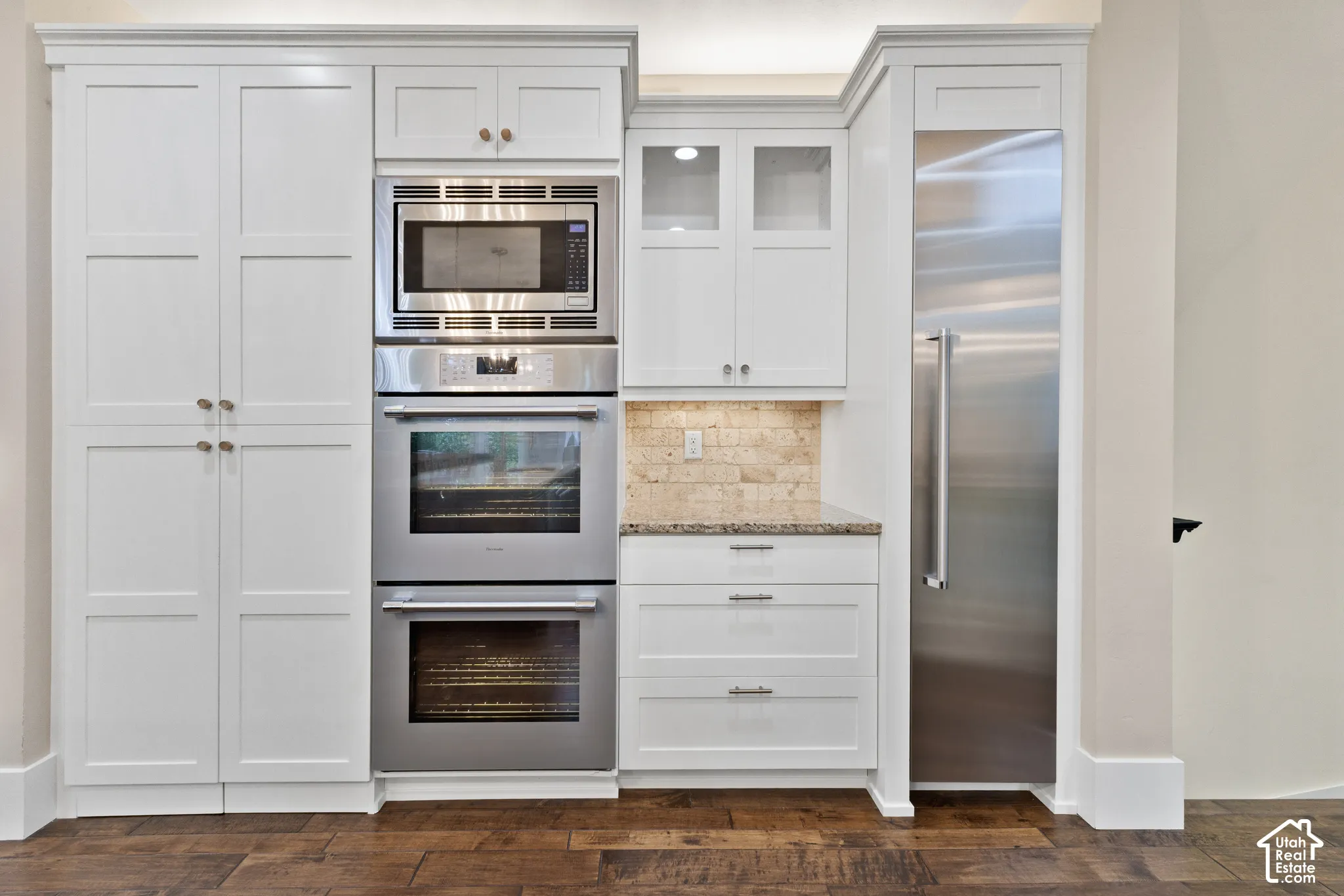 Custom cabinetry featuring shaker-style doors, integrated stainless steel refrigerator, built-in double ovens with microwave, stone-finish backsplash, and wood-finish flooring