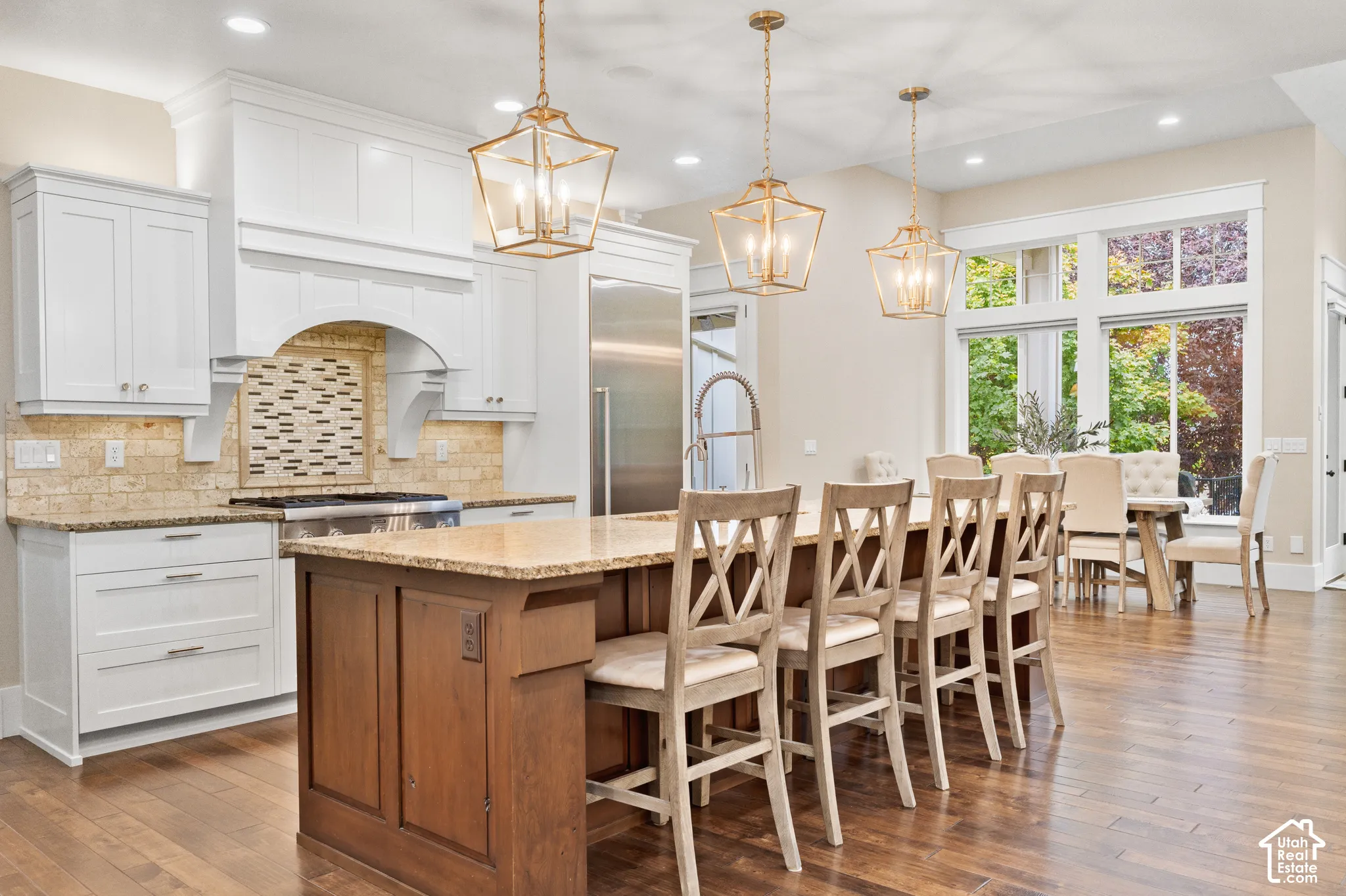 Kitchen featuring a spacious central island with stone countertop, white cabinetry with integrated range hood, wood-finish flooring, three gold-tone geometric pendant lights, and large paneled windows