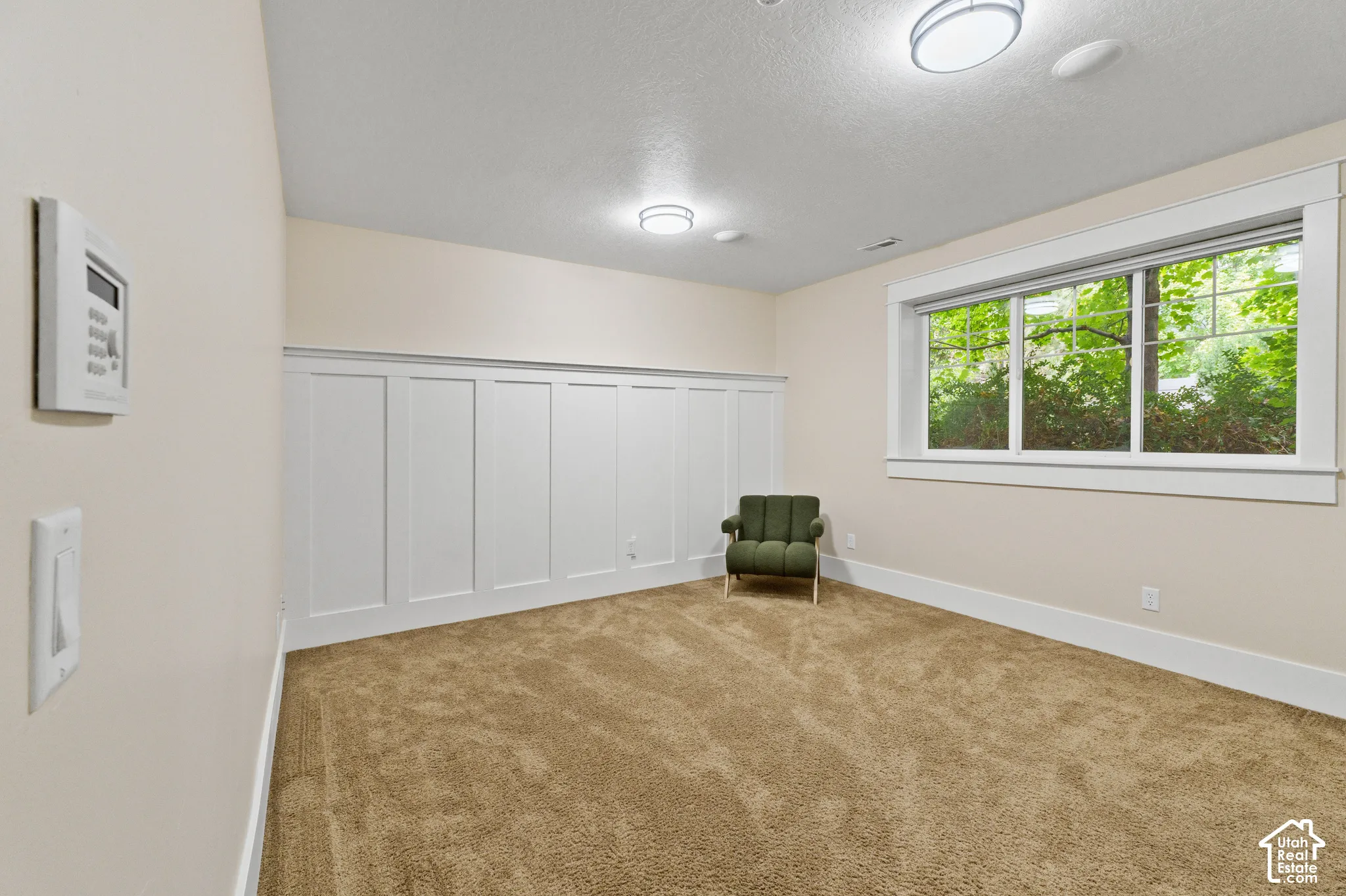 Interior room featuring white wainscoting, neutral carpeting, flush-mount ceiling lighting, and a large window with exterior greenery