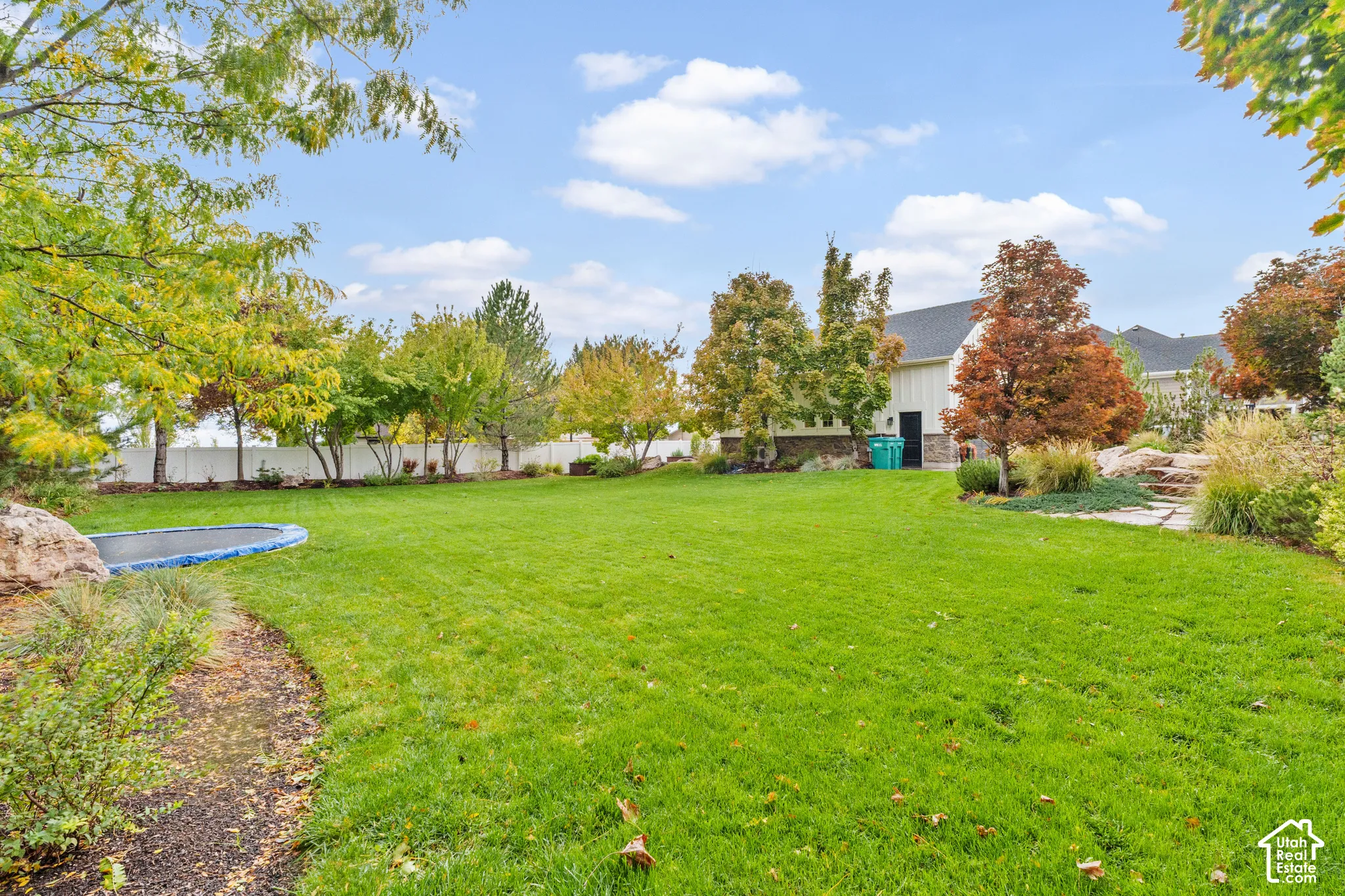 Expansive backyard featuring a manicured lawn, mature trees, stone landscaping, and a white privacy fence