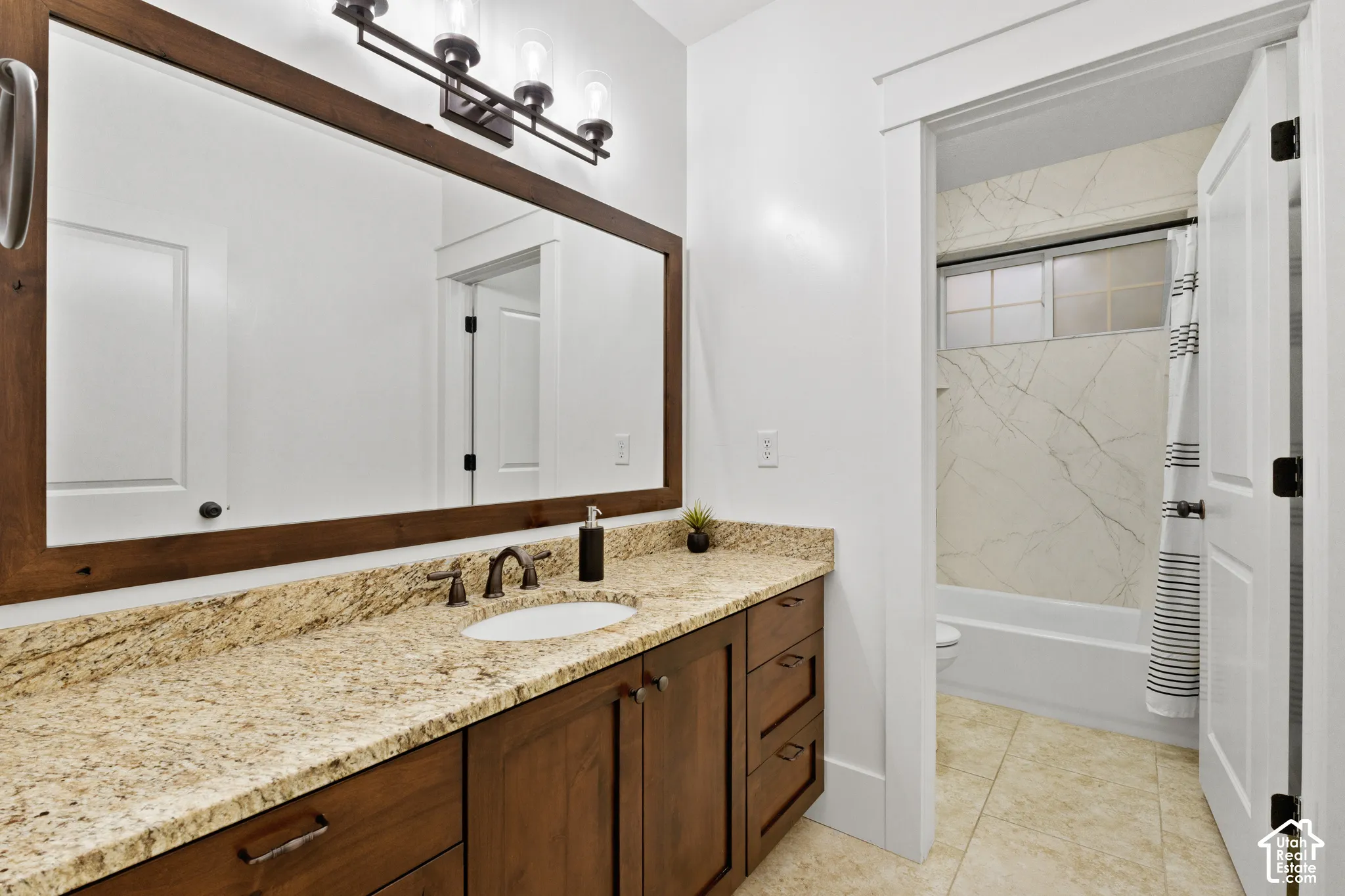 Stained wood cabinetry with stone-finish countertop, undermount sink, three-light fixture, framed vanity mirror, and bathtub enclosure with stone-finish wall surround
